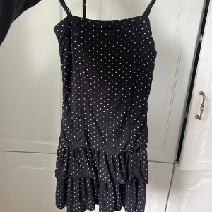 Svart prickig kort klänning med volang - Supersöt svart kort klänning med vita prickar och smala axelband. Klänningen har volang nertill som ger en lekfull känsla. Perfekt för dig som gillar en klassisk men trendig look. Materialet känns lätt och flowy.
