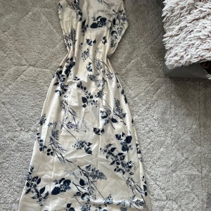 Lång blommig klänning med meshdetalj - Snygg långklänning i ljus satin med marinblått blommönster. Klänningen har tunna axelband och en unik meshdetalj i ryggen med korsade band. Perfekt för dig som vill sticka ut med en elegant och trendig look.