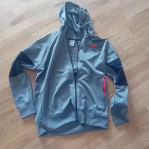 Nike Air Max grå vindjacka med huva - Nike Air Max vindjacka i grått med mörkare detaljer på ärmarna och rosa logga fram och bak. Jackan har huva, hel dragkedja och två fickor med rosa dragkedjor. Perfekt för dig som gillar sportig och clean stil.