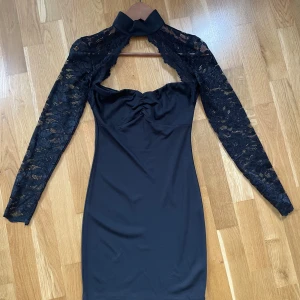 Svart spetsklänning Model Behaviour S - Supersnygg svart kortklänning från Model Behaviour med långa spetsärmar och hög krage. Klänningen har figurnära passform, axelbandslös byst och transparent spets upptill. Perfekt för dig som vill sticka ut med en elegant och edgy look.
