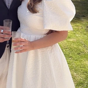 Vit kort klänning med puffärm - Superfin vit kort klänning med puffiga kortärmar och öppen rygg. Klänningen har en lätt struktur i tyget och markerad midja som ger en söt och trendig look. Perfekt för sommarens alla tillfällen!
