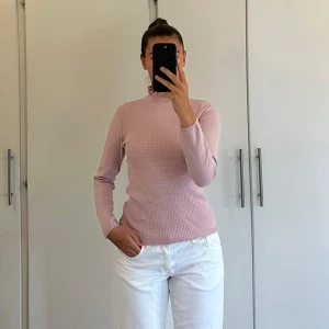 Ljusrosa stickad polotröja - Supersöt ljusrosa stickad polotröja med ribbad, volangig krage och långärmad passform. Tröjan har ett diskret strukturerat mönster framtill och sitter tajt men bekvämt. Perfekt för lager-på-lager och ger en fräsch vibe till din outfit.