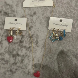 Set med örhängen och halsband hjärta/stjärna - Säljer ett färgglatt smyckeset från Fashion Jewellery med flera örhängen i guld, blått och rött, samt ett halsband med rött hjärta. Örhängena har detaljer som hjärta, stjärna, pärla och geometriska former. Perfekt för dig som gillar att mixa och matcha smycken. Aldrig använd.