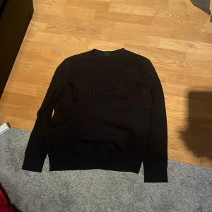 Svart BOSS sweatshirt i bomull M - Svart sweatshirt från BOSS i storlek M. Tröjan har en clean look med BOSS-logga i svart på bröstet och rund hals. Långa ärmar och ribbade muddar ger en snygg passform. Perfekt för dig som gillar stilrena och klassiska plagg.