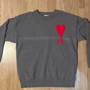 Ami stickad tröja med hjärta - Stickad tröja från Ami med ikoniskt rött eller vitt hjärta och bokstaven A på bröstet. Finns i grått, marinblått, vitt och svart. Klassisk rund halsringning och ribbade muddar. Perfekt för dig som vill ha en stilren men ändå unik look.