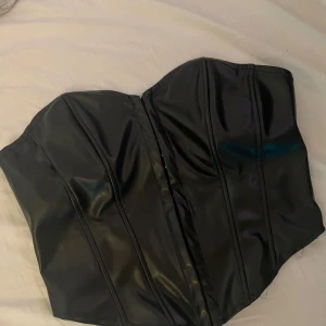 Svart korsett i skinnimitation - Säljer en svart korsett med snygg skinnlook och markerade sömmar. Toppen är axelbandslös och har hakknäppning framtill för en tight passform. Perfekt för dig som vill ha en edgy vibe och gillar statement-plagg.