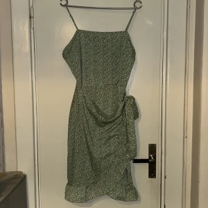 Grön blommig omlottklänning M - Supersöt grön omlottklänning med vita små blommor, smala axelband och volangkant nertill. Klänningen har knyt i sidan och är perfekt för sommarens alla tillfällen. Lätt och luftig känsla, passar dig som gillar romantisk stil.