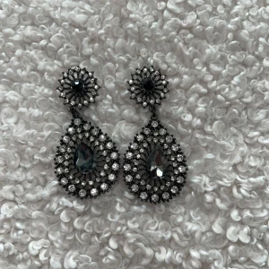 Svarta örhängen - Snygga stora örhängen i svart metall med droppformad design och massor av glittrande stenar. De har en cool vintagekänsla och är perfekta för att sticka ut. Stenarna är klara och svarta, och hela örhänget har en lyxig vibe med mycket bling.