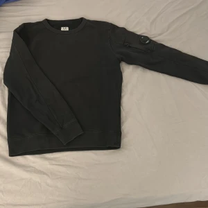 Svart sweatshirt från C.P. Company - Svart sweatshirt från C.P. Company med klassisk rund hals och lång ärm. Tröjan har den ikoniska linsen på ena ärmen och är tillverkad i 100% bomull för en skön och avslappnad känsla. Perfekt för dig som gillar streetwear med stilren touch. Det är 2 sprickor på själva märket.