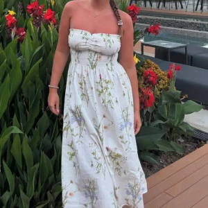 Vit blommig axelbandslös långklänning - Superfin vit axelbandslös långklänning med smock upptill och söt rosett vid bysten. är perfekt för varma dagar. Kommer med band till axlarna som man kan ta på och av 💐