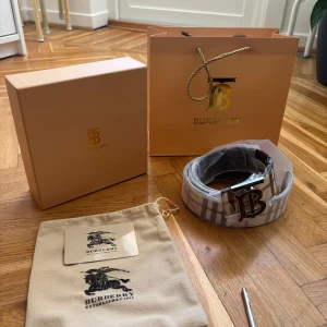 Burberry rutig skärp med B-spänne - Exklusivt Burberry-bälte med klassiskt rutigt mönster i beige, svart och vinrött. Snyggt metallspänne med stor B-logga framtill. Bältet är tillverkat i skinn och har Burberrys ikoniska rutor på båda sidor, en ljus och en mörk. Perfekt accessoar för att lyfta din outfit.
