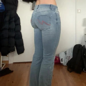 Ljusblå bootcut jeans med broderi - Säljer ett par ljusblå jeans med bootcut passform och snyggt rött broderi på bakfickorna. Jeansen har låg midja och klassisk femficksdesign. Perfekta för dig som gillar en avslappnad men ändå trendig look.