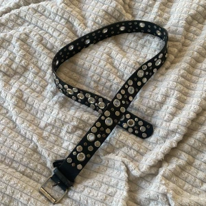 Svart bälte med silvriga nitar - Coolt svart bälte i skinn med massor av silvriga nitar och hål i olika storlekar längs hela bandet. Bältet har en klassisk metallspänne och ger en edgy vibe till vilken outfit som helst. Perfekt för dig som vill sticka ut med detaljer.