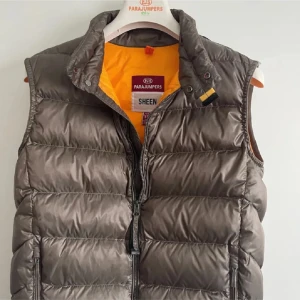 Brun dunväst från Parajumpers - Snygg brun dunväst från Parajumpers med quiltad design och gul insida. Västen har hög krage, dragkedja framtill och två fickor med dragkedja. Perfekt att slänga över en hoodie för en chill look. Klassisk Parajumpers-detalj vid halsen.