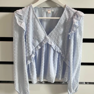 Ljusblå volangblus från Gina Tricot - Supersöt ljusblå blus från Gina Tricot med volangdetaljer och prickigt strukturmönster. Blusen har v-ringning, långa ärmar med resår vid handlederna och volanger längs axlar och framtill. 