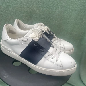 Valentino Garavani Open sneakers vit/blå - Snygga Valentino Garavani Open sneakers i vitt skinn med bred mörkblå läderdetalj över snörningen och ikoniska nitar baktill. Klassisk rund tå, platt sula och vita skosnören. Perfekta för dig som gillar lyxiga och stilrena sneakers.