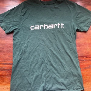 Grön Carhartt t-shirt med logga - Mörkgrön t-shirt från Carhartt med klassisk vit logga tryckt på bröstet. Modellen är enkel med rund hals och korta ärmar, perfekt för en avslappnad streetwear-look. Tillverkad i mjuk bomull som känns skön mot huden.