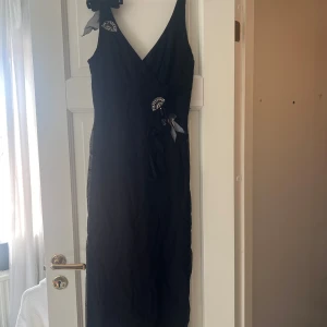 Svart långklänning med rosetter och pärlor - Unik svart långklänning i mesh med omlott-skärning, v-ringad hals och dekorativa rosetter. Klänningen har pärlbroderade detaljer och en stor mesh-rosett på axeln. Perfekt för dig som vill sticka ut med coola detaljer och elegant vibe.