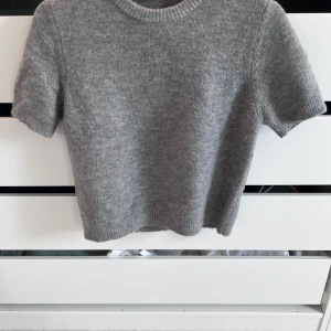 Grå stickad kortärmad tröja Zara - Grå stickad tröja från Zara i storlek M men känns som S. Tröjan har korta ärmar, rund halsringning och ribbade kanter. Perfekt för lager-på-lager och chill vibes. Enkel att matcha med både jeans och kjol.