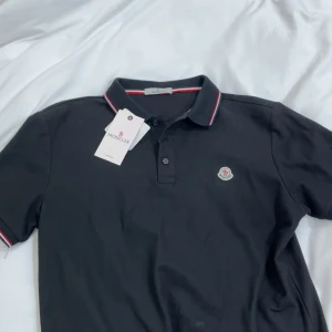 Moncler piketröja - Svart pikétröja från Moncler med klassisk krage och knäppning framtill. Tröjan har Moncler-logga på bröstet och snygga rödvita detaljer på krage och ärmslut. Tillverkad i bomull för en skön och stilren känsla.