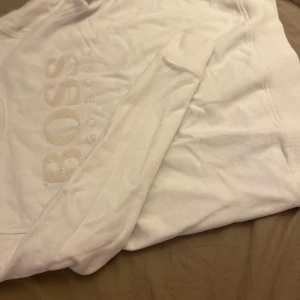 Vit sweatshirt från Hugo Boss - Snygg vit sweatshirt från Hugo Boss med broderad logga framtill. Tröjan har rund hals, långa ärmar och ribbade muddar vid ärmslut och nederkant. Perfekt för en clean och stilren look.