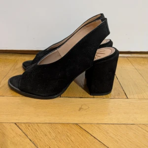 Svarta äkta mocka klackskor/slingbacks från K.Cobler - Stilrena svarta pumps i äkta mocka från K.Cobler med öppen tå och bred klack. Skorna har en snygg slingback-design med elastiskt band bak för extra komfort. Knappt använda, se bilder. Perfekta för dig som vill ha en klassisk men trendig look.