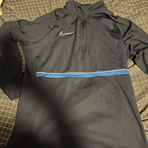 Nike Dri-Fit långärmad träningströja - Nike Dri-Fit långärmad träningströja i mörkblått med halv dragkedja och vit Nike-logga på bröstet. Snygg blå rand över bröstet och sportig passform. Perfekt för träning eller chill dagar. Materialet är polyester som andas och håller dig torr.