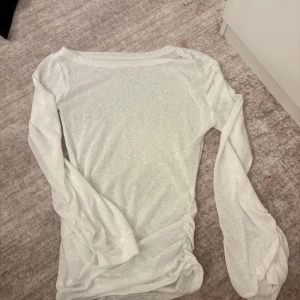 Vit långärmad topp med rynkade detaljer - Vit långärmad topp i tunn bomullsmix med rynkade detaljer längs ena sidan och vid ärmsluten. Modellen har en enkel, rund halsringning och är lätt transparent. Perfekt för dig som gillar en clean och stilren look med en twist.