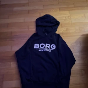 Svart hoodie från BORG Sportwear - Svart hoodie från BORG Sportwear med stor vit logga framtill. Klassisk modell med huva och dragsko, samt ribbade muddar vid ärmslut och nederkant. Perfekt för en sportig och avslappnad stil.