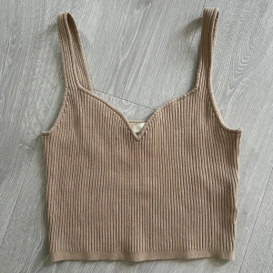 Beige ribbad croppad linnetopp - Säljer en beige ribbad linnetopp med breda axelband och v-ringad front. Toppen är croppad och har en snygg, enkel design som passar perfekt till jeans eller kjol. Materialet är mjukt och stretchigt för en bekväm passform.