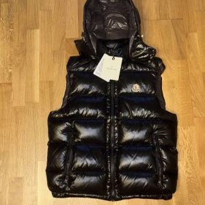 Svart dunväst från Moncler med huva - Säljer en svart, glansig dunväst från Moncler med avtagbar huva och två dragkedjefickor framtill. Västen har quiltad design och klassisk Moncler-logga på bröstet. Perfekt för lager-på-lager och riktigt snygg till streetwear eller casual outfits.