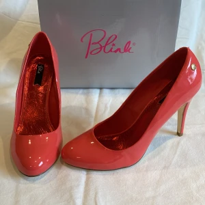 Korallröda pumps från Blink, storlek 41 - Snygga korallröda pumps från Blink i glansigt lackmaterial. Skorna har rund tå, 11cm hög klack och guldfärgade detaljer på hälen. Perfekta för dig som vill sticka ut med färg och stil. Levereras med extra klacktoppar och i orginalkartong. Aldrig använda, endast provade.