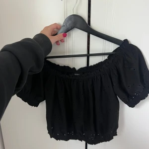 Svart offshoulder blus med broderi - Supersöt svart offshoulder blus med kort puffärm och broderade hålmönster. Croppad modell med resår i både halsringning, ärmslut och nederkant för en snygg passform. Perfekt för varma dagar och en trendig look.