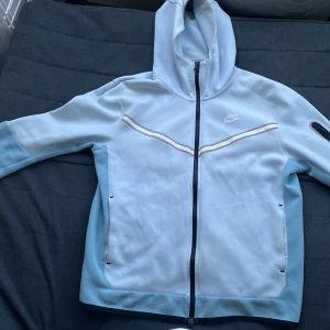 Nike Tech ljusblå med luva M - Nike vindjacka i ljusblått med hel dragkedja, huva och coola detaljer som ficka med dragkedja på ärmen. Jackan har vita och beige ränder över bröstet och är perfekt för dig som gillar sportig stil. Snygg passform och klassisk Nike-logga på bröstet.