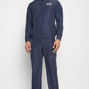 EA7 mörkblå träningsdräkt, storlek S - En snygg träningsdräkt från EA7 Emporio Armani. Dräkten är i en mörkblå färg och har en sportig design med logotypen på bröstet och armen.   Den är knappt använd och är i nyskick!