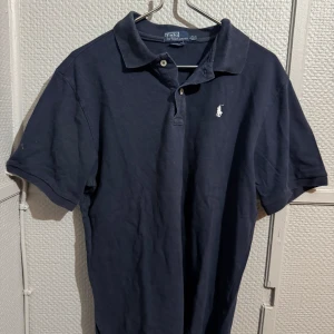 Mörkblå pikétröja från Ralph Lauren - Klassisk mörkblå pikétröja från Ralph Lauren med vit broderad logga på bröstet. Tröjan har korta ärmar, krage och knäppning med två knappar. Tillverkad i bomull och har en snygg, tidlös look som funkar till allt.