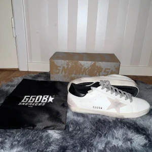 Golden goose skor  - Snygga vita GGDB sneakers med grå stjärna på sidan, grå snörning och svart hälparti. Skorna har en klassisk låg profil och är tillverkade i skinn med detaljer i mocka. Perfekta för dig som gillar en stilren men ändå trendig look.