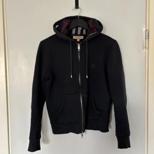 Svart hoodie från Burberry XS - Svart hoodie från Burberry med dragkedja, huva med klassiskt rutigt foder och snörning. Broderad logga på bröstet och två fickor framtill. Perfekt för dig som vill ha en stilren och lyxig streetwear-look.