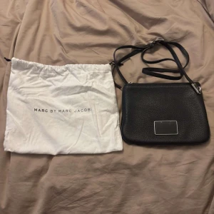 Svart axelväska Marc by Marc Jacobs - Snygg svart axelväska från Marc by Marc Jacobs i äkta skinn med silverfärgad dragkedja och metallplakett framtill. Stilren och enkel design med justerbar axelrem. Kommer med original dustbag. Perfekt storlek för det viktigaste.