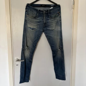 Blå Replay jeans med slitningar - Säljer ett par blå Replay jeans med snygga slitningar och råa benslut. Jeansen har klassisk femficksdesign, ljus tvätt och detaljerade bakfickor med kontrastsömmar. Märkespatch i läder baktill och dragkedja framtill. Perfekta för en avslappnad streetstil.