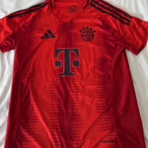 FC Bayern München Adidas matchtröja röd S - Röd FC Bayern München fotbollströja från Adidas med svarta detaljer och klubbmärke på bröstet. Tröjan har korta ärmar, V-ringning och klassiska Adidas-ränder på axlarna. Tillverkad i lätt och ventilerande material, perfekt för träning eller match.