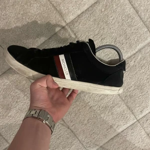 Moncler skor  - Snygga svarta sneakers från Moncler med klassisk vit sula och röd-vit rand på sidan. Skorna har snörning och rund tå, samt Moncler-logga på plösen. Perfekta för dig som vill ha en stilren och exklusiv look.