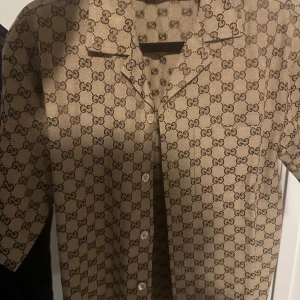 Beige Gucci skjorta med GG-mönster - Snygg beige skjorta från Gucci med klassiskt GG-monogram över hela plagget. Skjortan har korta ärmar, knappar framtill och är tillverkad i en lyxig blandning av linne och polyester. Perfekt för dig som vill sticka ut med en ikonisk designerlook. Storleken passar för dem som bär mellan S-M