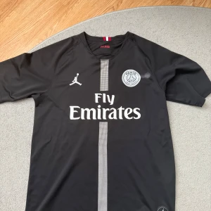 PSG x Jordan svart matchtröja - Svart PSG fotbollströja i samarbete med Jordan, med vit logga och emblem på bröstet samt Fly Emirates-tryck. Tröjan har korta ärmar, diskret randig detalj framtill och är tillverkad i lätt, ventilerande material. Perfekt för dig som älskar streetstyle och fotboll. SNABB AFFÄR = BÄTTRE PRIS