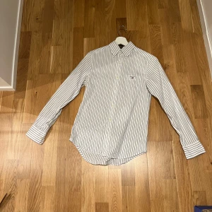 Vit och blå randig skjorta från Gant - Snygg skjorta från Gant i vitt med tunna blå ränder. Klassisk krage, långärmad och med knappar framtill samt på manschetterna. Skjortan har en bröstficka och är tillverkad i mjuk bomull. Perfekt för en clean och stilren look.