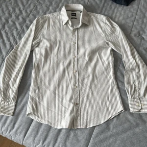 Randig skjorta från BOSS i bomull - Snygg randig skjorta från BOSS i vitt och grått. Skjortan har klassisk krage, långa ärmar med manschett och knäppning framtill. Casual fit med stretch för extra komfort. Perfekt för dig som gillar stilren och enkel design.