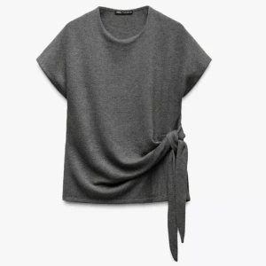 Grå knytblus från Zara - Supermjuk grå blus från Zara med korta ärmar och snygg knytning i sidan. Toppen har rund halsringning och är gjord i ett stickat material som känns både chill och stilren. Perfekt att styla med jeans eller kjol för en clean look.