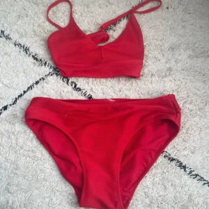 Röd ribbad bikini med korsade band - Snygg röd bikini med ribbad struktur och korsade axelband i ryggen. Överdelen har en sportig form med smala axelband och byxan är klassisk med medelhög midja. Perfekt för stranden eller poolen i sommar!