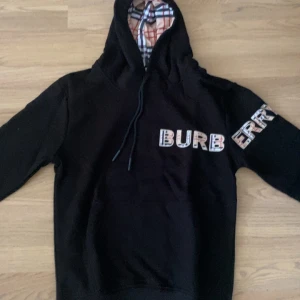 Svart Burberry hoodie med rutiga detaljer - Svart hoodie från Burberry med en luva. Märket är skriven med   stora bokstäver med rutigt mönster på bröstet och ärmen. Tröjan har snörning i luvan och en avslappnad passform. Perfekt för dig som vill ha en svart tröja som passar med alla olika färgbyxor. Använd i 3 gånger , men ville byta min stil. 
