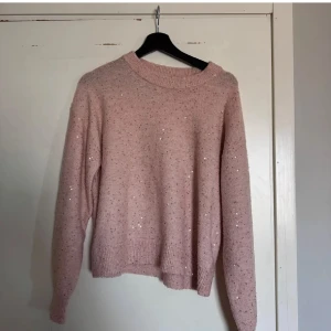 Rosa stickad tröja med paljetter - Superfin ljusrosa stickad tröja med paljettdetaljer som ger ett subtilt glittrande intryck. Tröjan har en rund halsringning, ribbade muddar och lång ärm. Perfekt för dig som vill ha något mysigt men ändå lite extra.